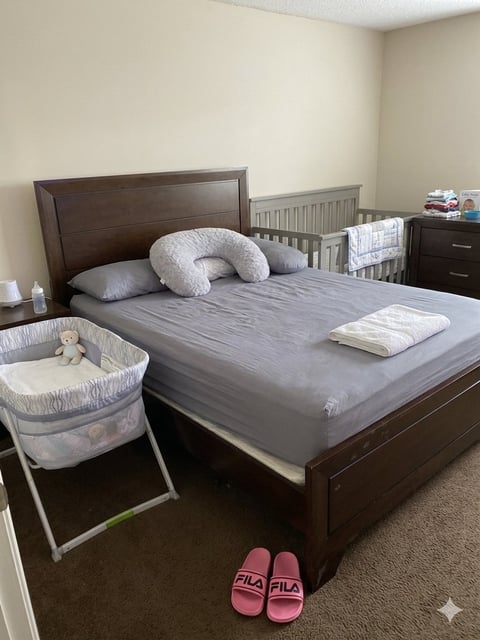 Master bedroom with dark wood bed, gray sheets, white nursing pillow, portable bassinet beside bed, pink slippers on carpeted floor, and crib in background