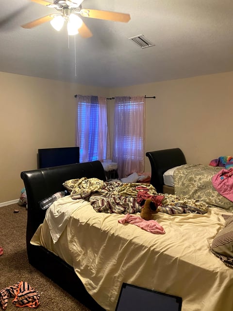 Bedroom with two black beds with rumpled bedding, ceiling fan, windows with blue curtains, and carpet floor
