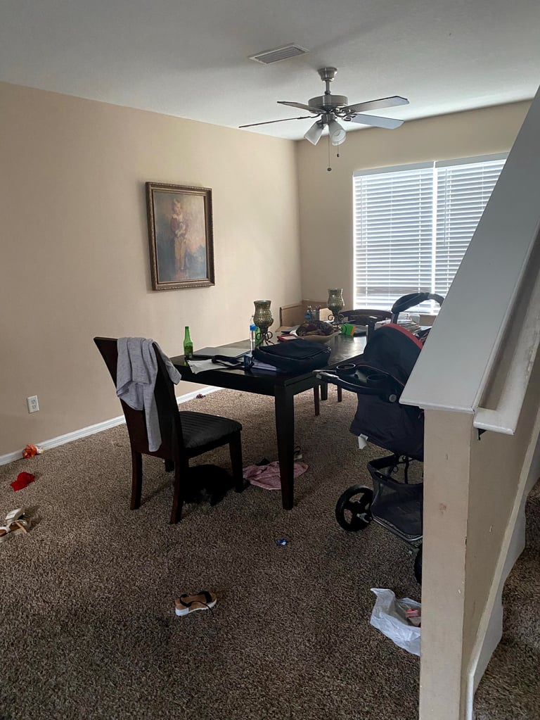 Messy home office with black desk, two chairs, ceiling fan, windows with blinds, and scattered items on carpeted floor