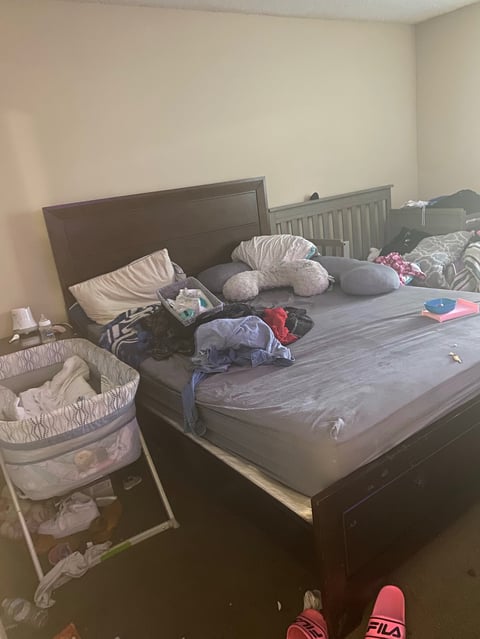 Messy bedroom with unmade bed covered in clothes and pillows, a portable crib, and various items scattered on the floor
