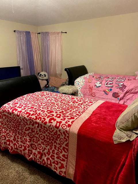 A bedroom with two beds covered in red and pink patterned bedding, sheer curtains on the window, and a black couch against the wall.