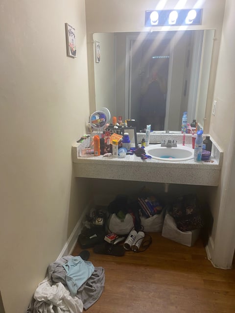 Cluttered bathroom vanity with mirror, cosmetics, and skincare products on countertop above storage cubbies filled with bags and clothing items on wooden floor