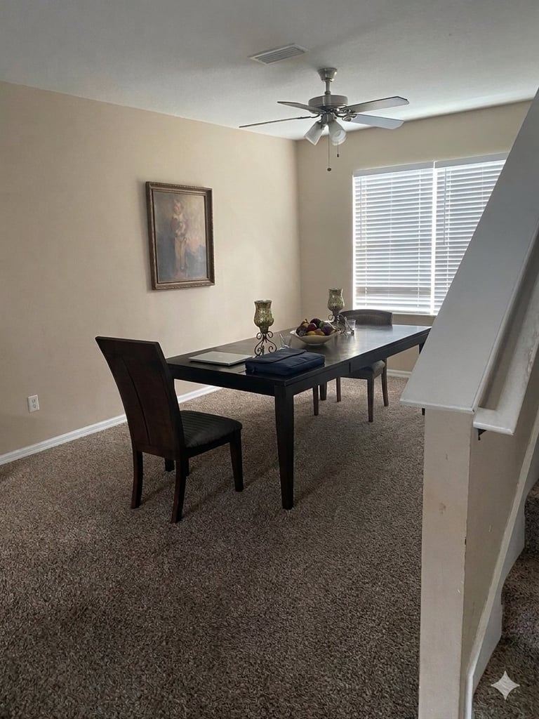 Modern home office with black desk, office chair, ceiling fan, and large window with blinds overlooking beige walls and carpet flooring